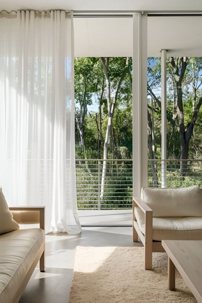 Bright, airy living room with floor-to-ceiling windows showcasing a forest view. White sheer curtains and minimalist furniture