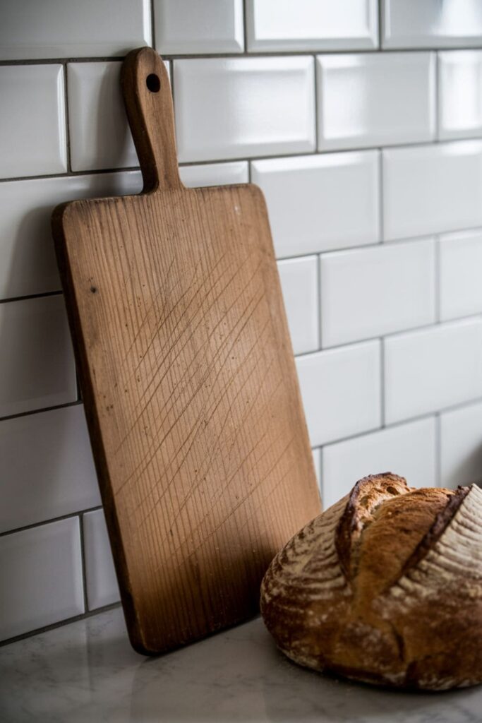 Large wooden breadboard leaning against a backsplash with a loaf of bread nearby